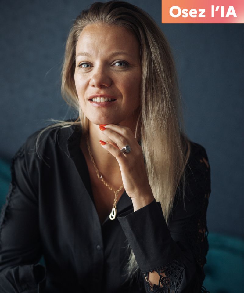 Portrait d'une femme avec des cheveux longs et blonds, portant une chemise noire, souriante et posant avec une main sur son menton. En haut à droite, le texte 'Osez l'IA' est affiché.