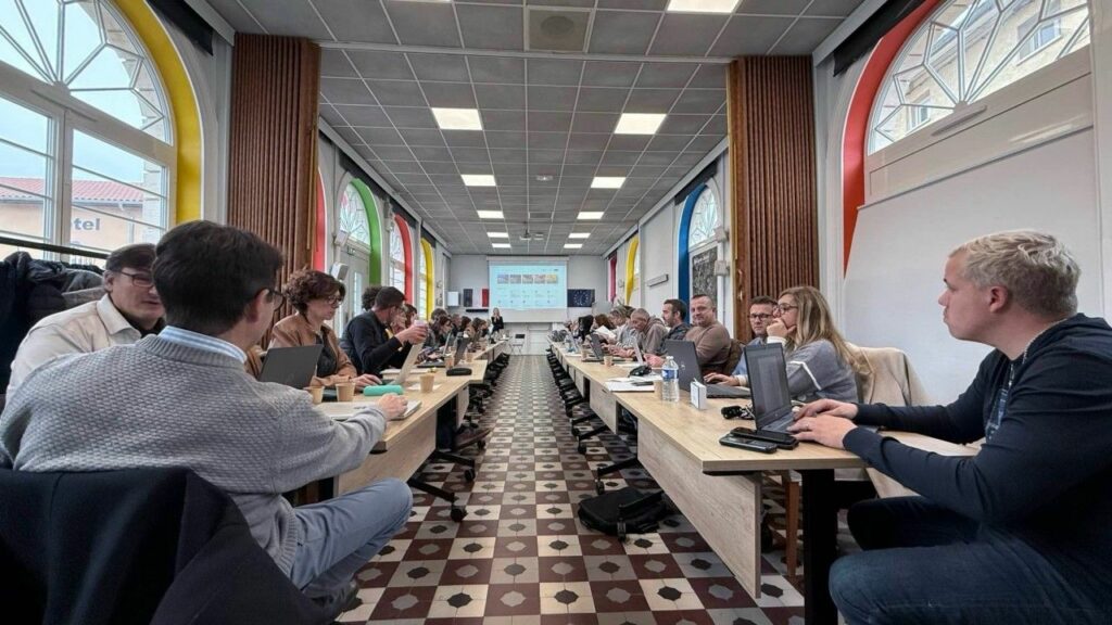 Une salle de formation avec des participants assis à des tables, utilisant des ordinateurs portables. À l'avant, un écran affiche des informations. Les fenêtres ont des rideaux colorés.